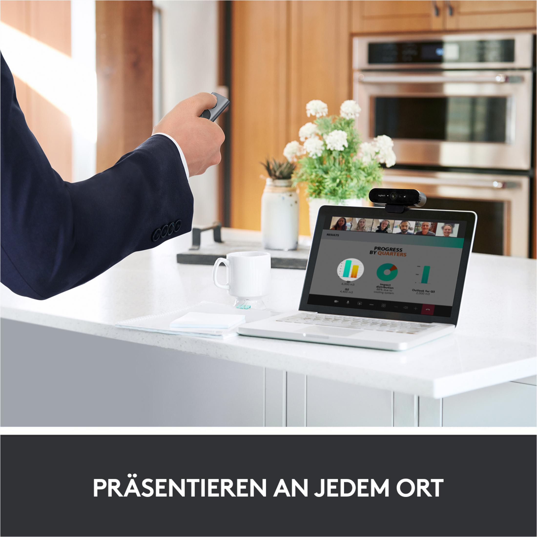 A man is presenting with a remote, looking at a laptop on a counter with a webcam attached.