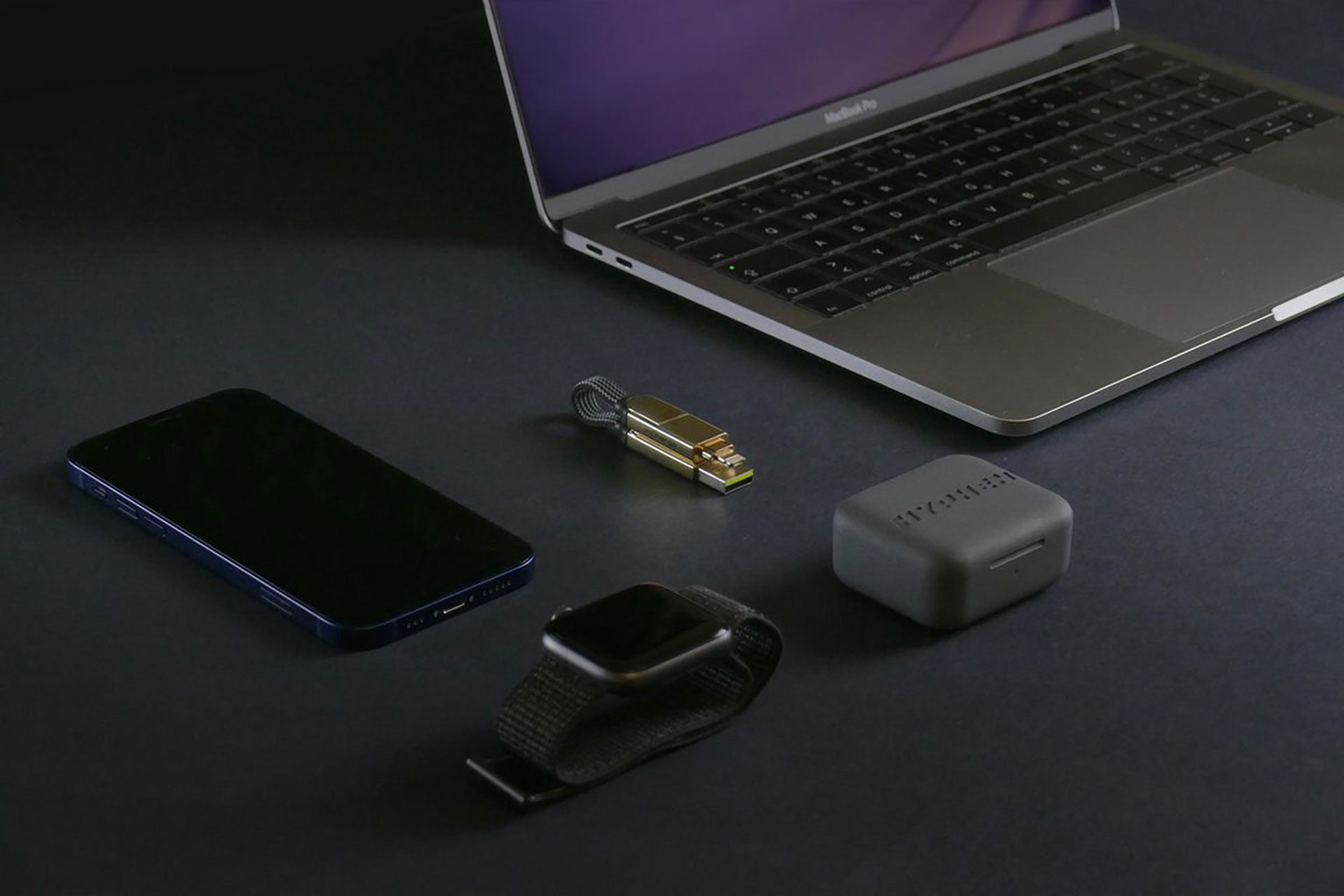 Un ordinateur portable, un téléphone, une smartwatch, une clé USB et un boîtier de chargement sont sur une surface sombre.