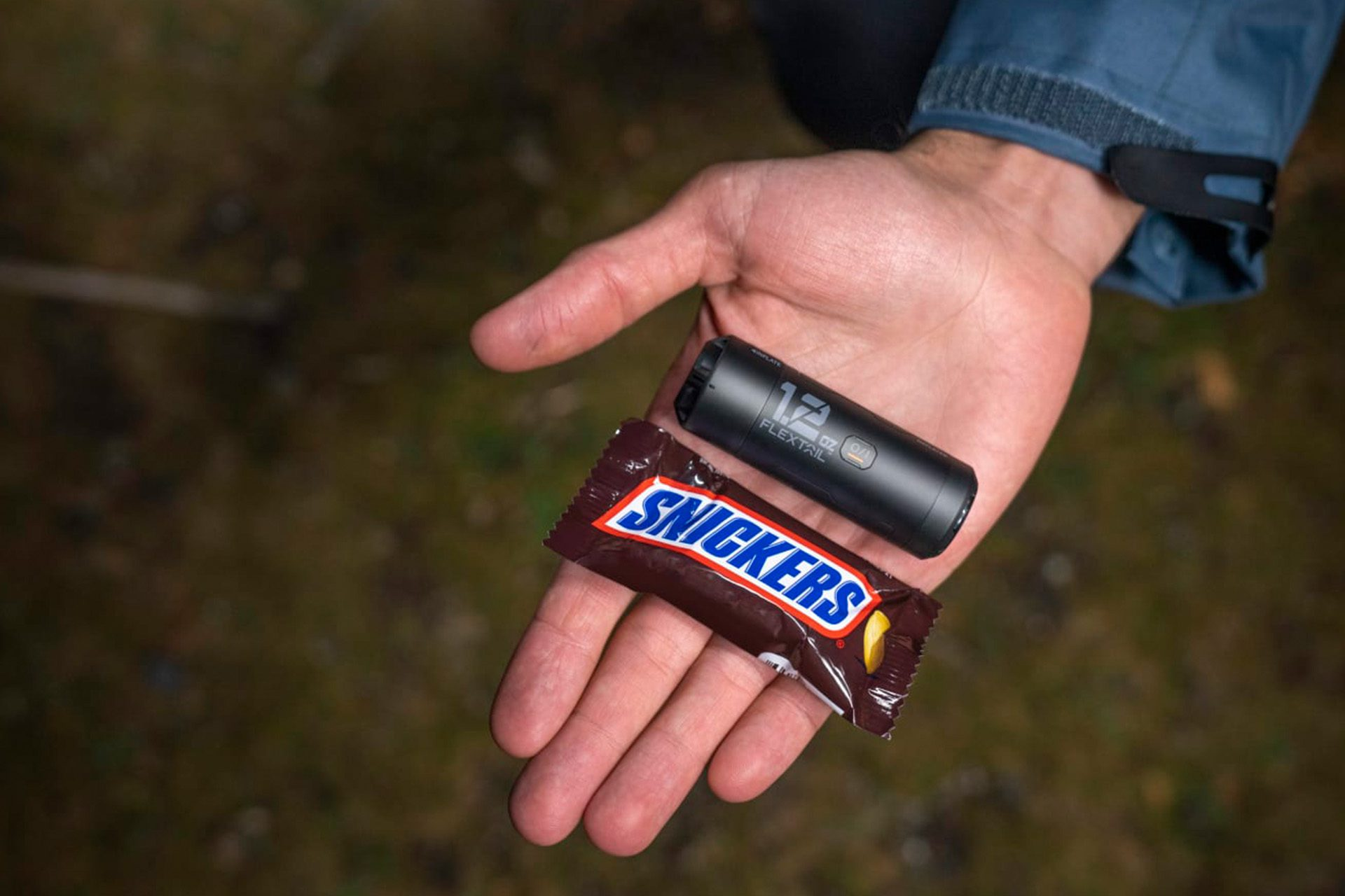 Hand hält Snickers und ein schwarzes FLECTAIL-Gerät. Der Hintergrund ist unscharf, wahrscheinlich im Freien.