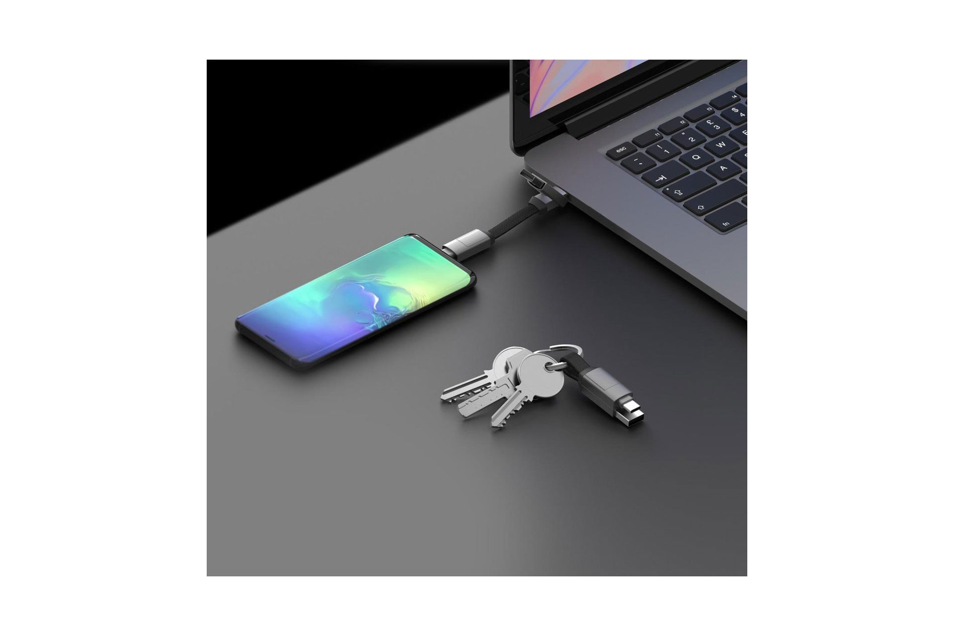 Een telefoon aangesloten op een laptop met sleutels ernaast op een zwart oppervlak. De laptop is open.