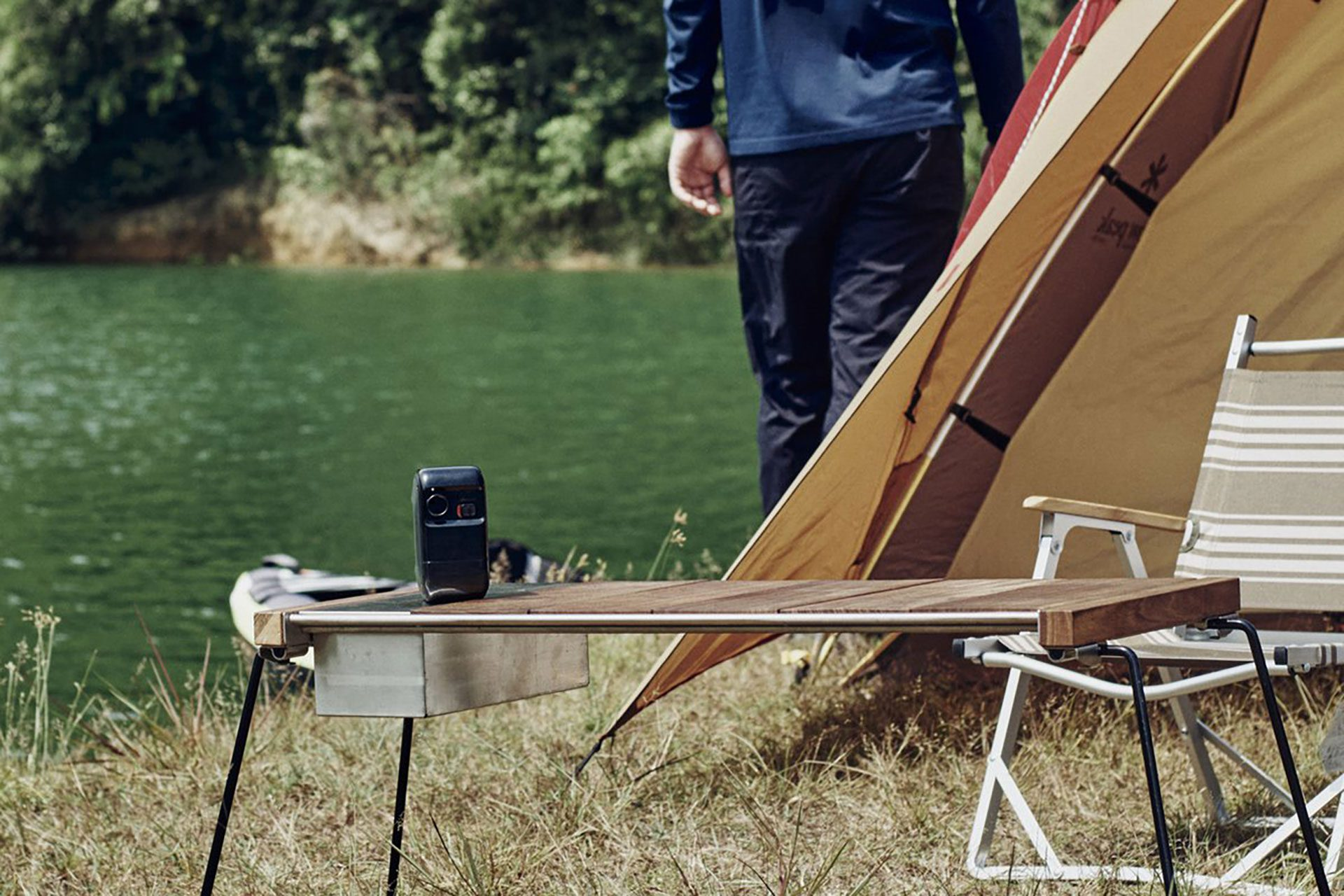 Camping-Szene mit Zelt, Tisch und Person am See. Die Szene ist im Freien.