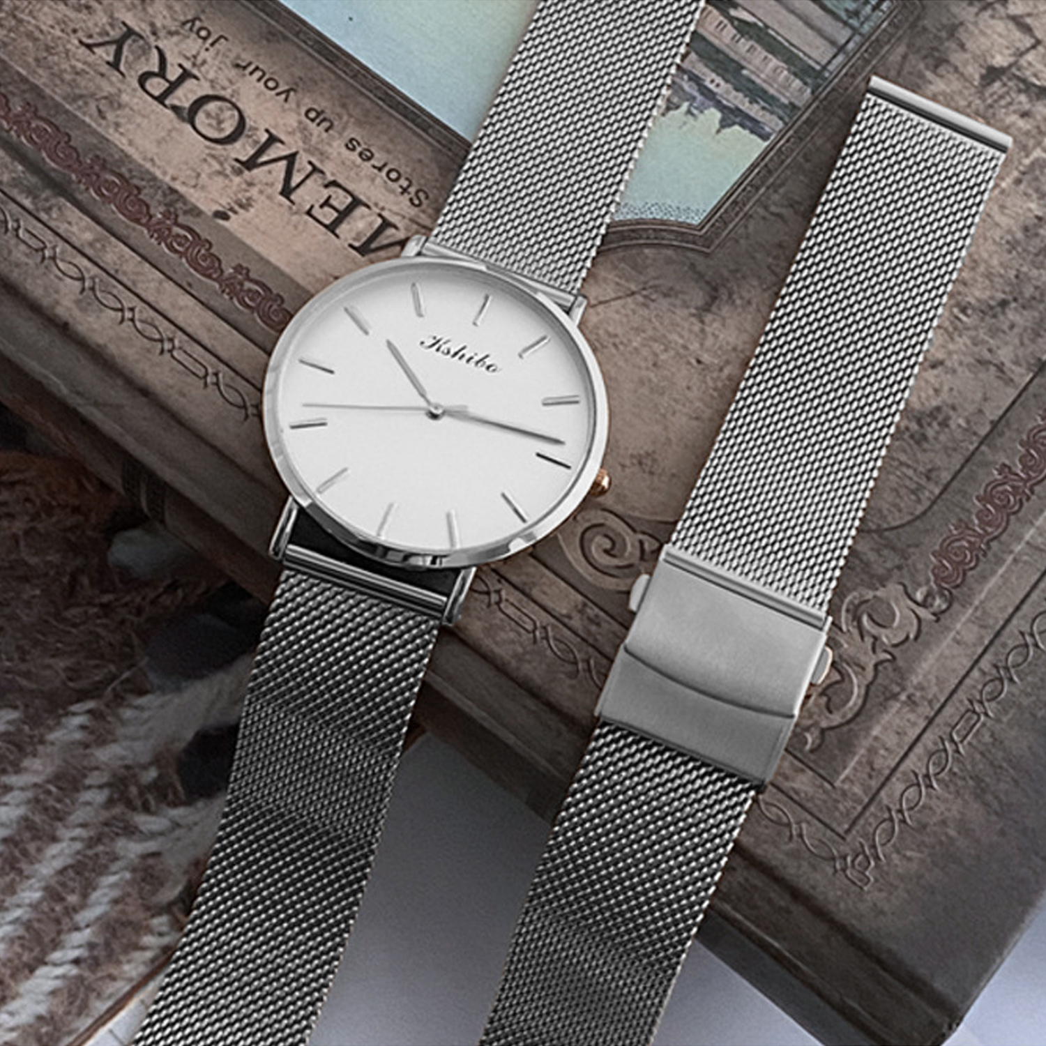 Srebrny zegarek na rękę z białą tarczą. Srebrna bransoleta. Zegarek i bransoleta leżą na książce. Neutralne tło.