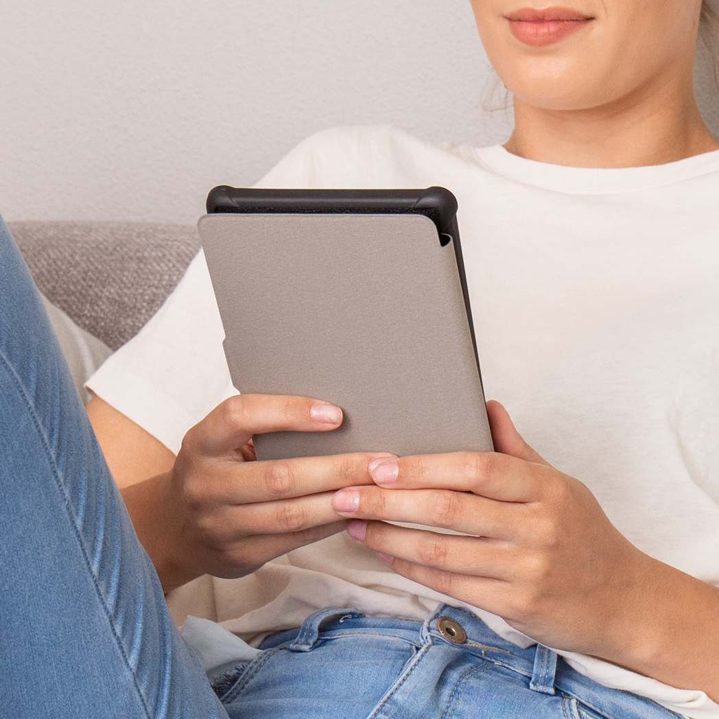 Persoon met tablet met grijze hoes. Wit t-shirt en blauwe jeans. Bank op de achtergrond.