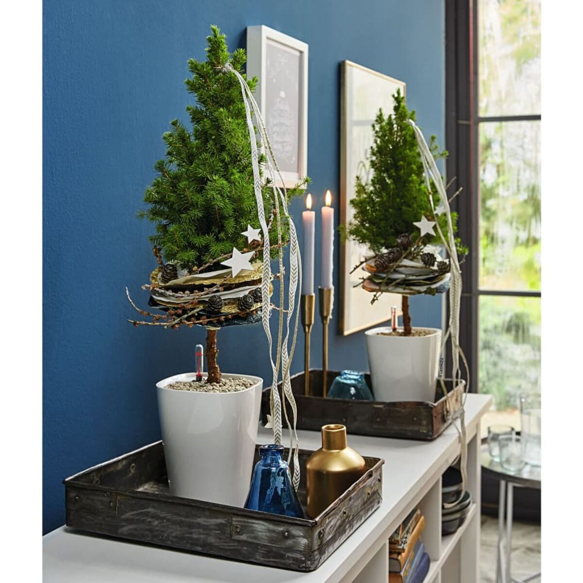Deux petits arbres de Noël sur une étagère blanche, avec des décorations et des bougies sur un fond de mur bleu.