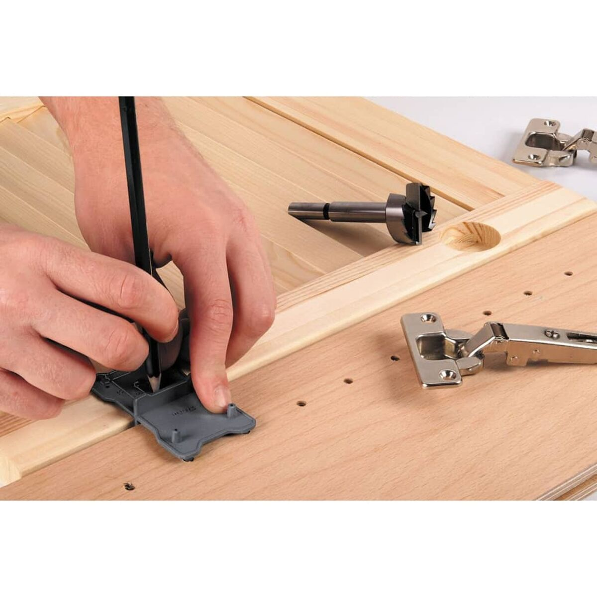 Person markiert mit einem Bleistift ein Holzbrett mit einer Schablone. Ein Bohrer und ein Scharnier liegen daneben.