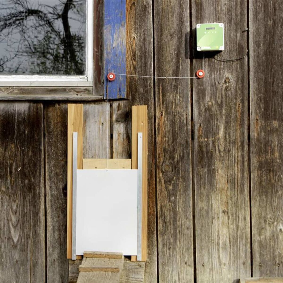Eine Holztür eines Hühnerstalls an einer verwitterten Holzwand. Eine Steuerbox ist oben montiert.