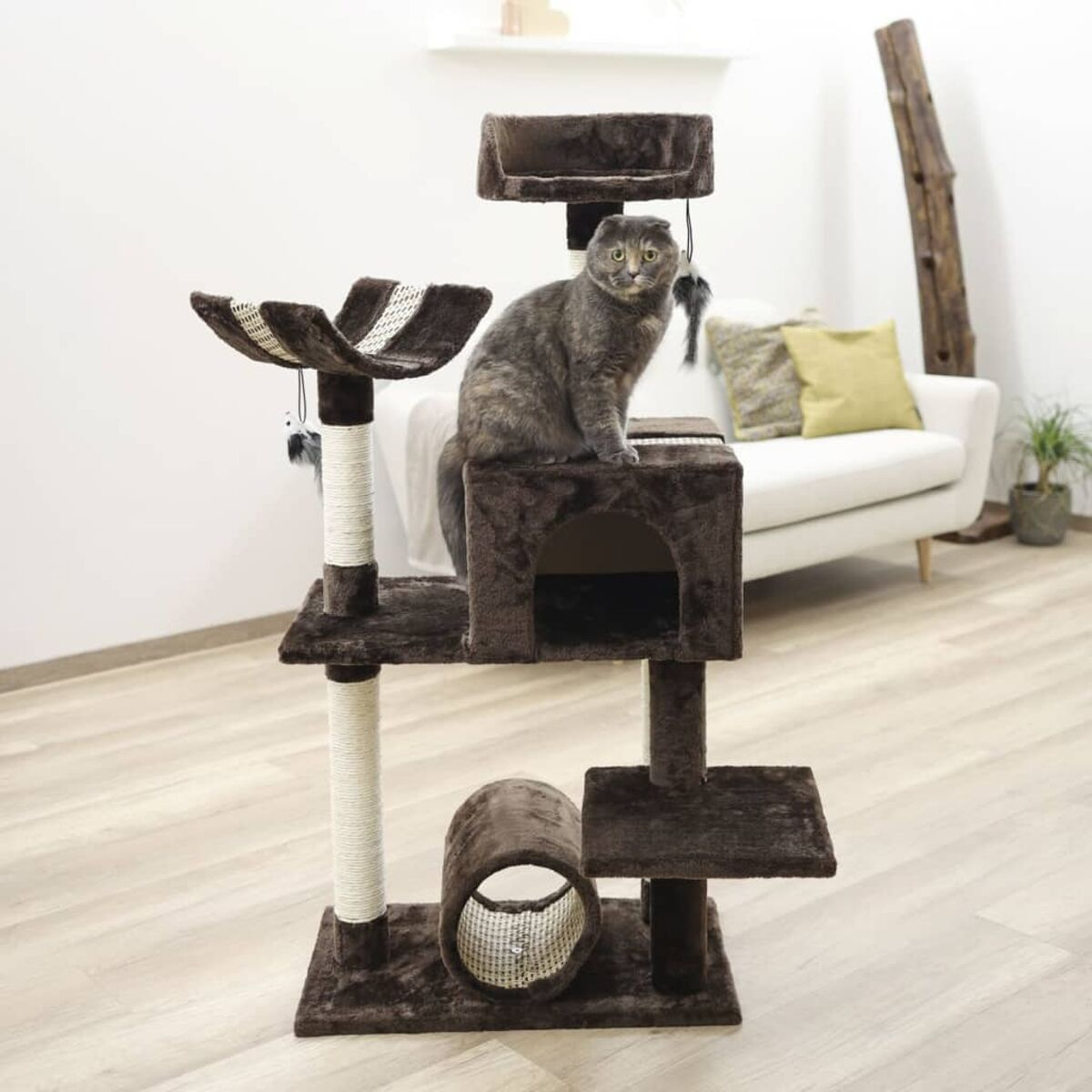 Eine graue Katze sitzt auf einem braunen Katzenbaum mit mehreren Plattformen und einem Tunnel. Im Hintergrund eine weiße Couch.