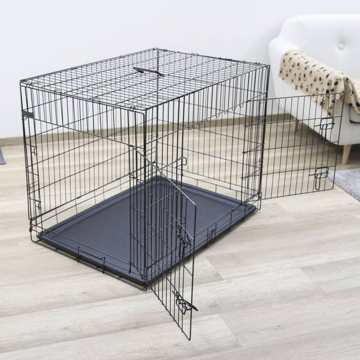 Schwarze Draht-Hundebox, teilweise geöffnet. Tablett innen. Holzboden, weißer Wandhintergrund.