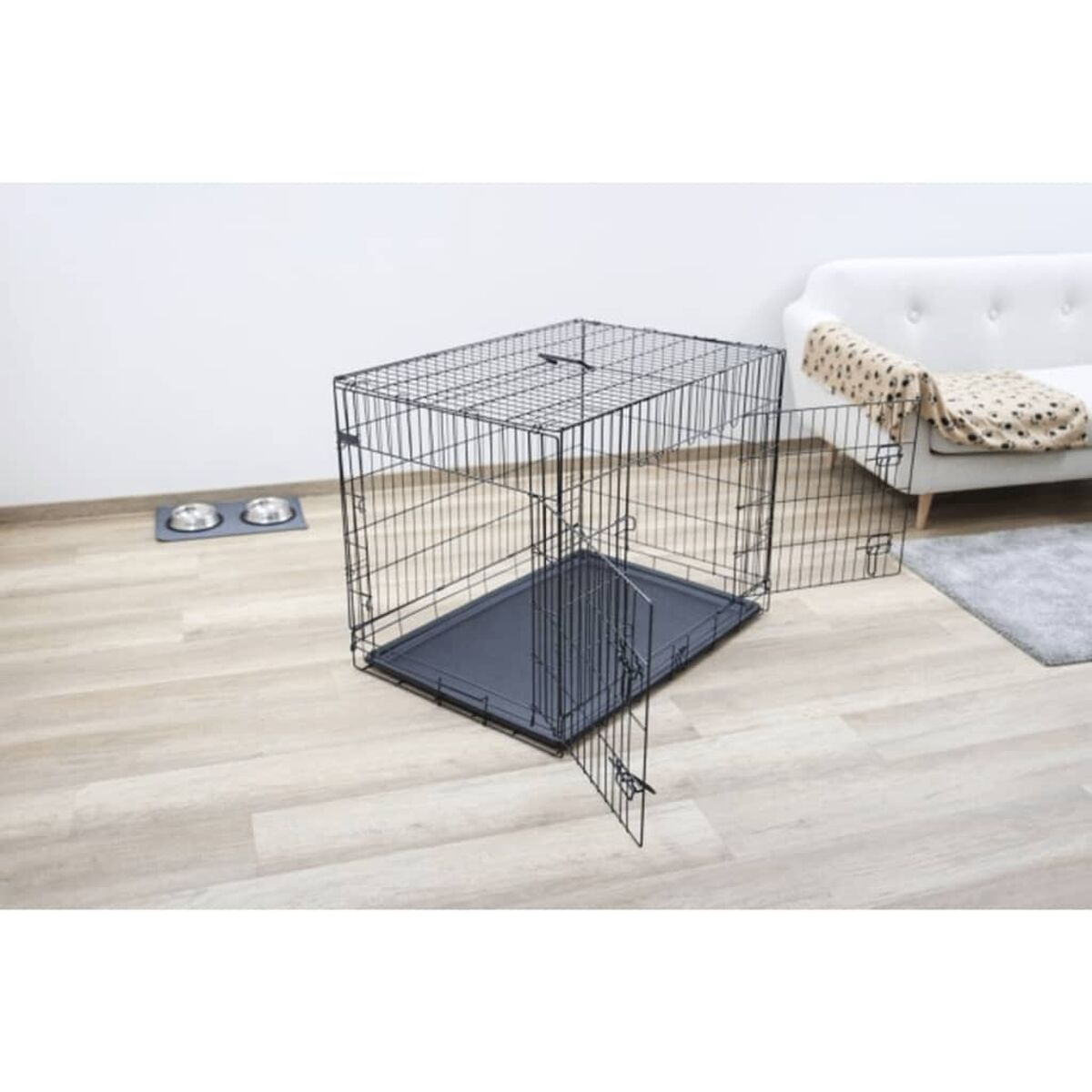 Schwarze Draht-Haustierbox, offene Tür. Holzboden. Näpfe im Hintergrund. Weißes Sofa.
