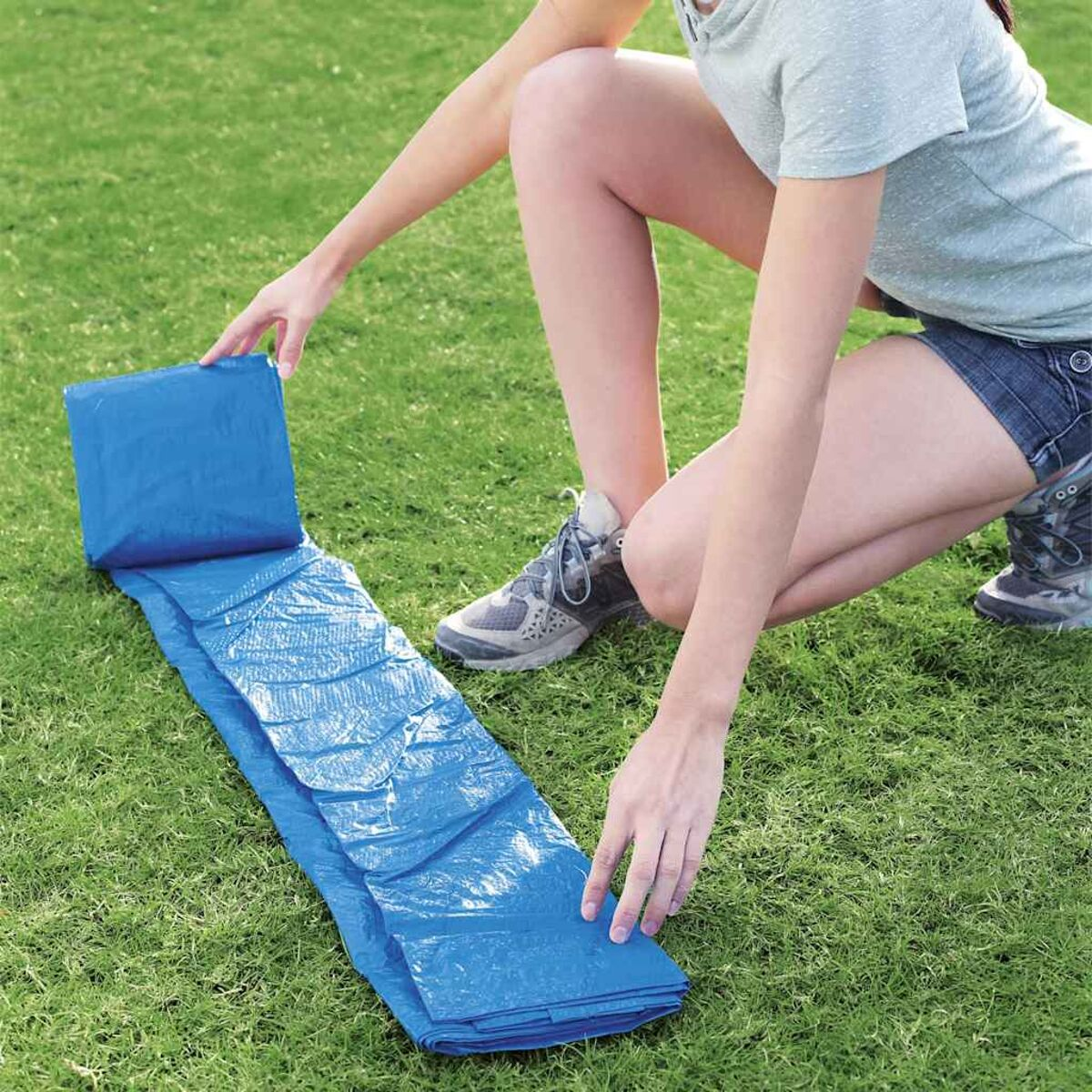 Person kniet und entfaltet eine blaue Poolmatte auf grünem Gras. Die Matte ist gefaltet.