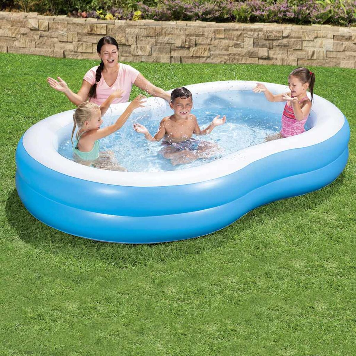 Vier Kinder und ein Erwachsener in einem aufblasbaren blauen Pool auf grünem Gras, spritzen Wasser.