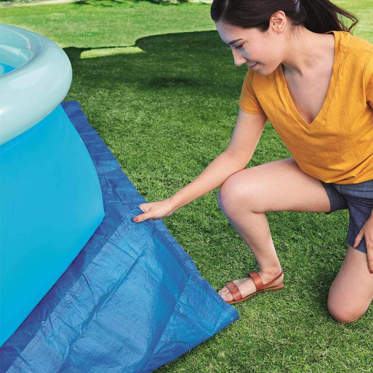 Frau kniet auf Gras, berührt eine blaue Plane neben einem teilweise sichtbaren Pool mit hellblauem Wasser.