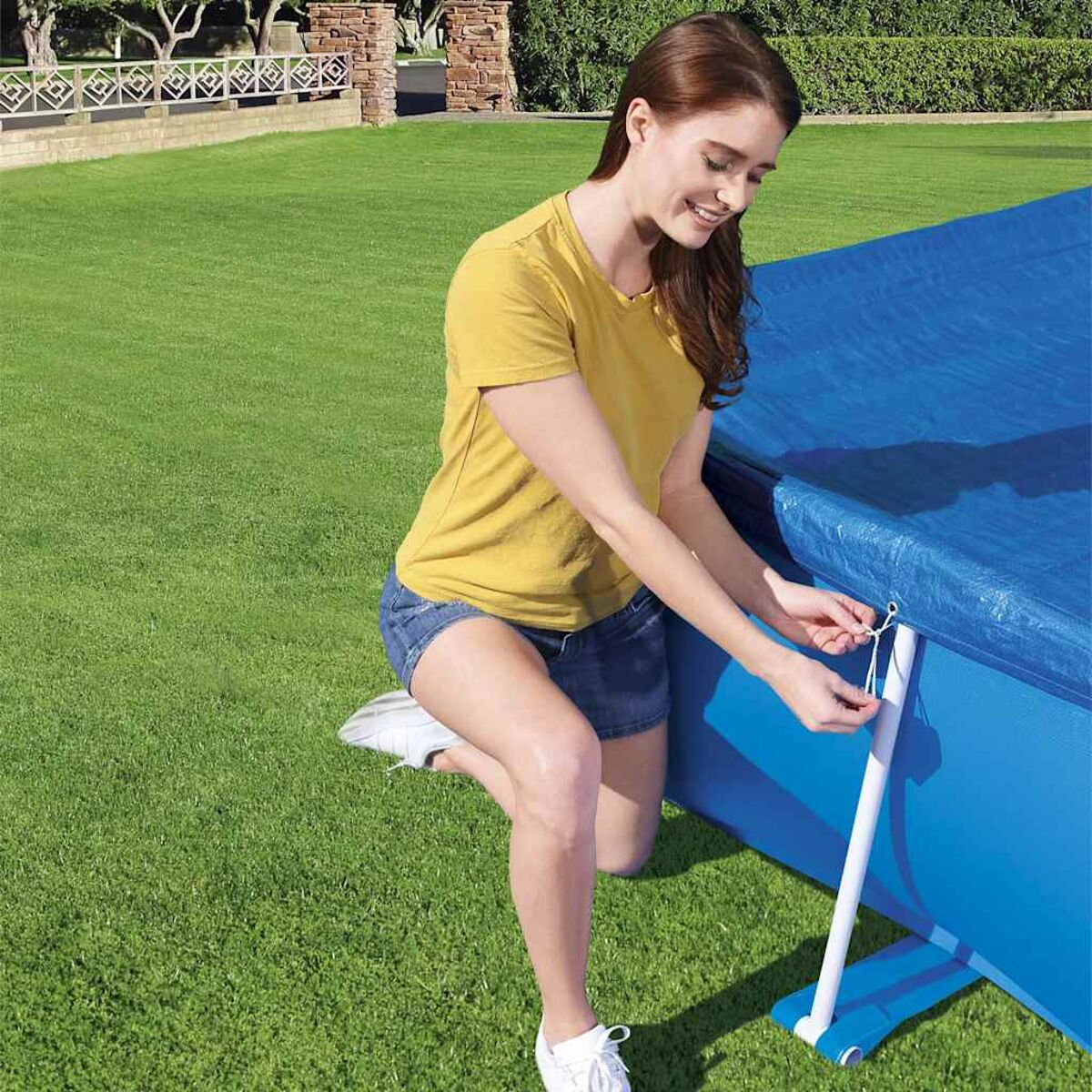 Frau in Shorts kniet und eine blaue Poolabdeckung auf grünem Gras befestigt.