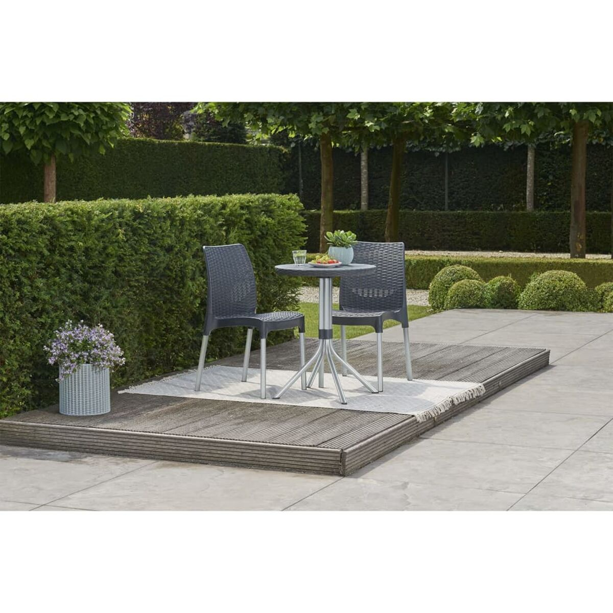 Terrassenmöbel-Set mit zwei Stühlen und einem kleinen Tisch. Ein Blumentopf und etwas Grün ist ebenfalls sichtbar.