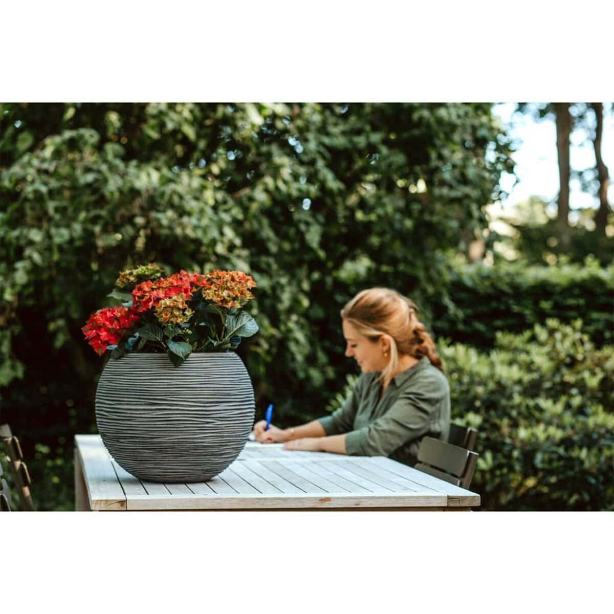 Frau draußen an einem Tisch beim Schreiben. Eine graue Vase mit roten Blumen steht auf dem Tisch. Grünes Laub im Hintergrund.