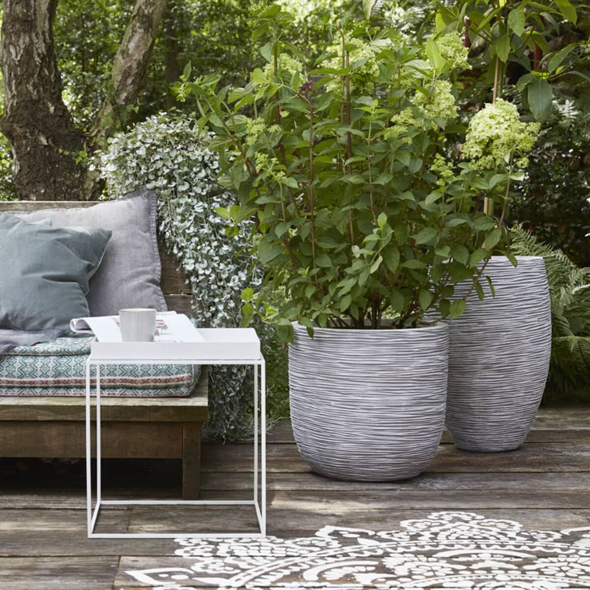 Eine Gartenszene mit Topfpflanzen auf einer Holzterrasse, einschließlich Tisch, Bank und Pflanzen.