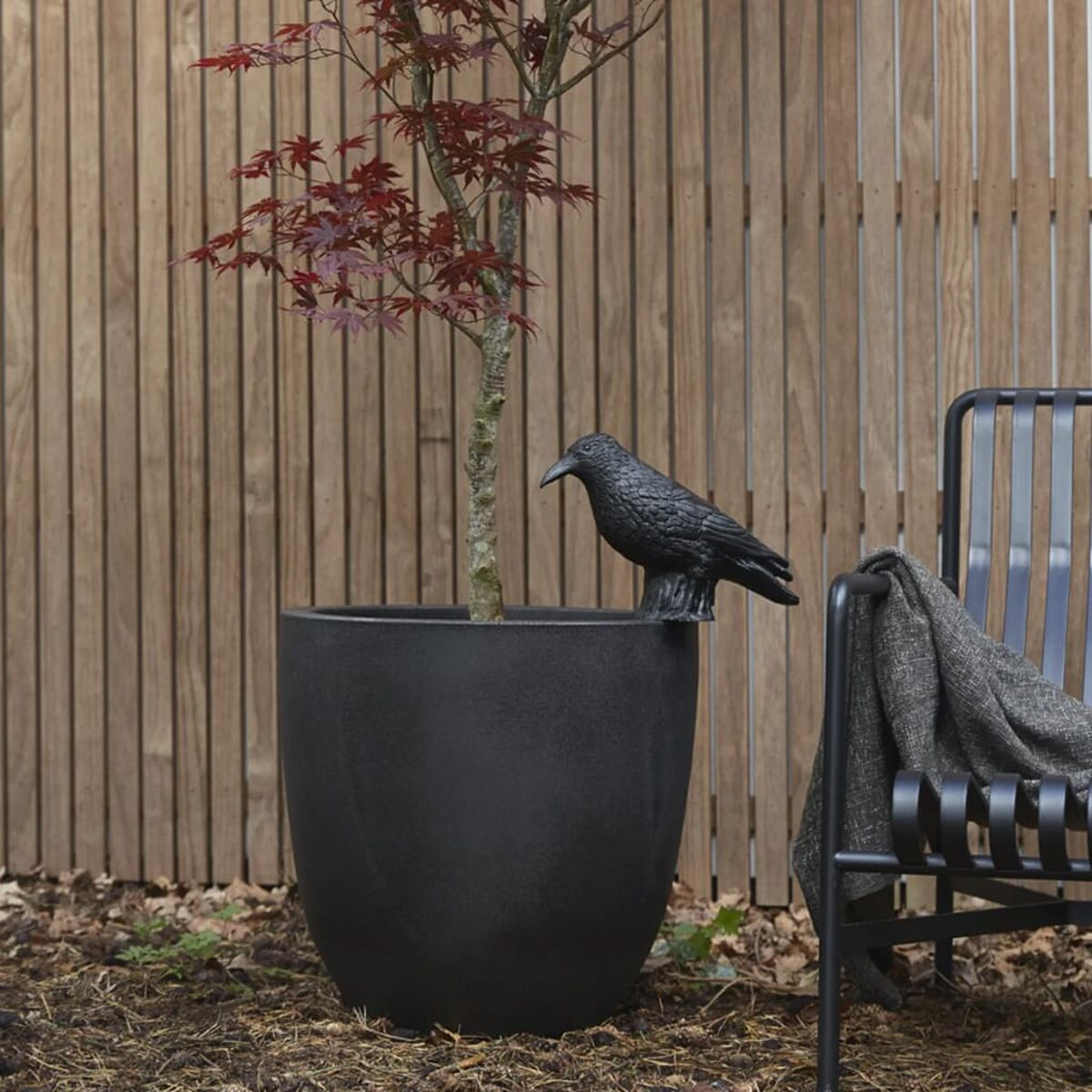 Ein Baum mit roten Blättern, eine schwarze Krähenstatue und ein Metallstuhl.