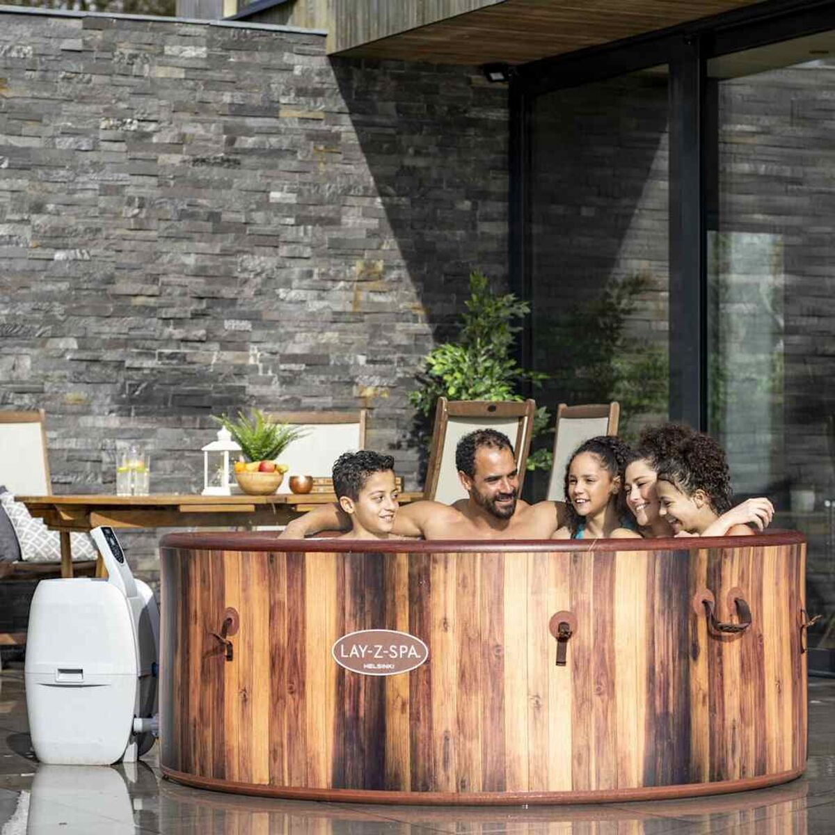 Familie in einem Whirlpool aus Holz. Steinmauer und Haus im Hintergrund.