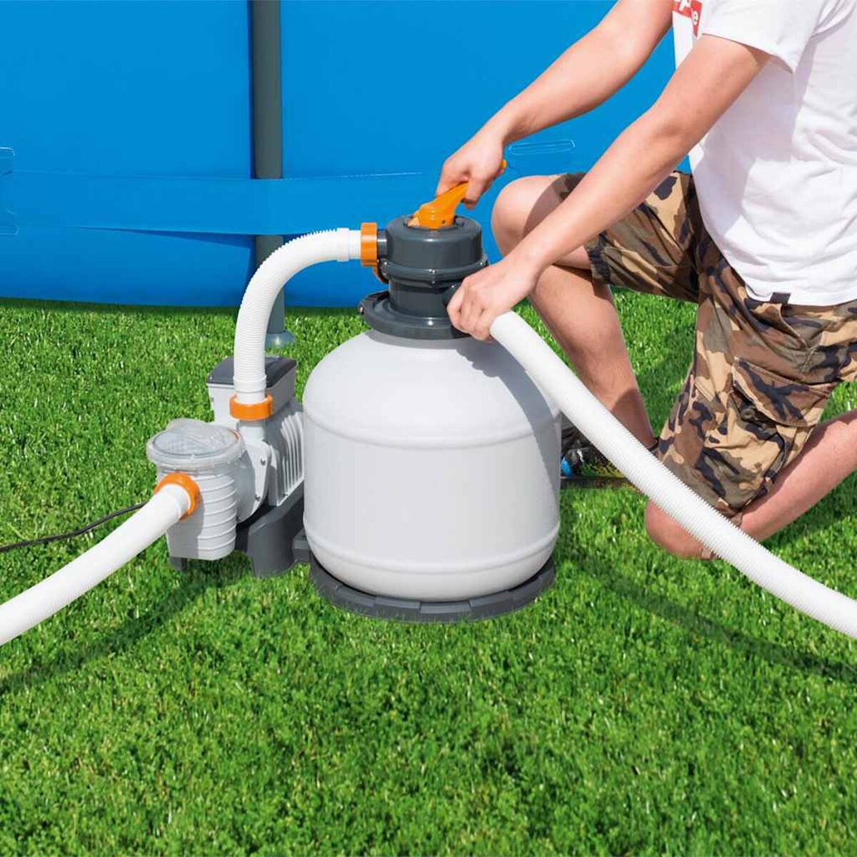 Eine Person bedient einen Poolfilter mit weißen Schläuchen auf grünem Gras.