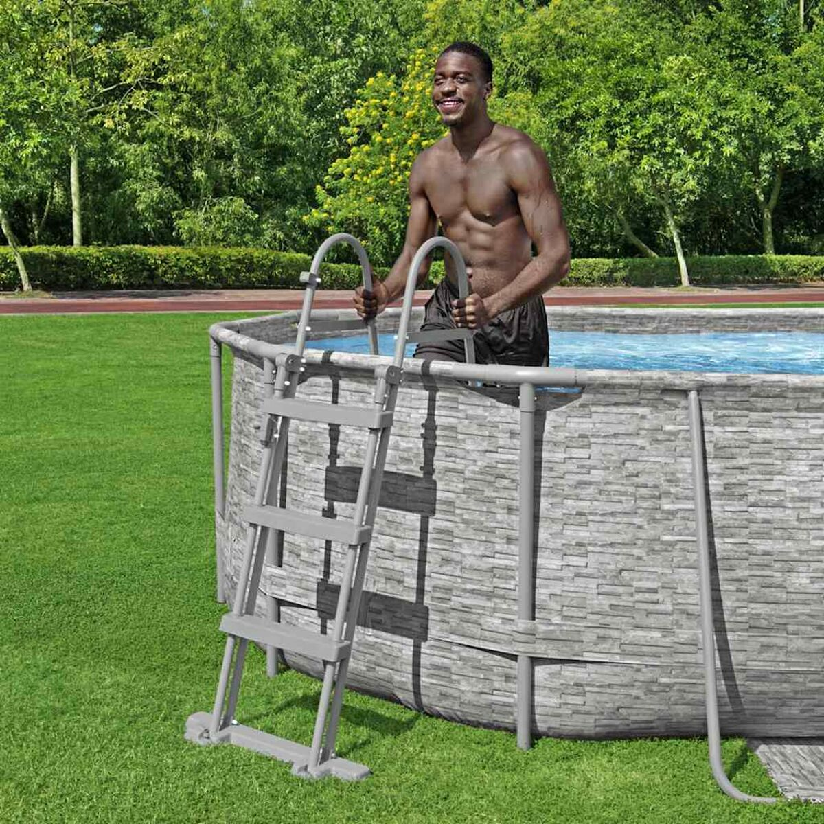 Mann im Pool, klettert über eine Leiter heraus. Grünes Gras und Bäume im Hintergrund. Pool hat steinförmige Außenseite.
