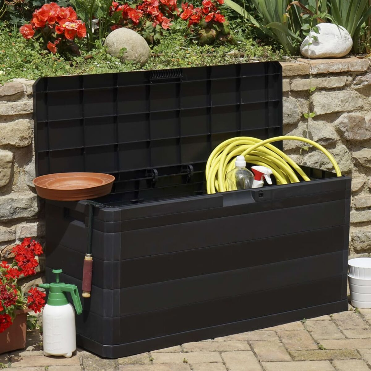 Schwarze Aufbewahrungsbox offen, Gartengeräte innen, rote Blumen und Steinmauer.