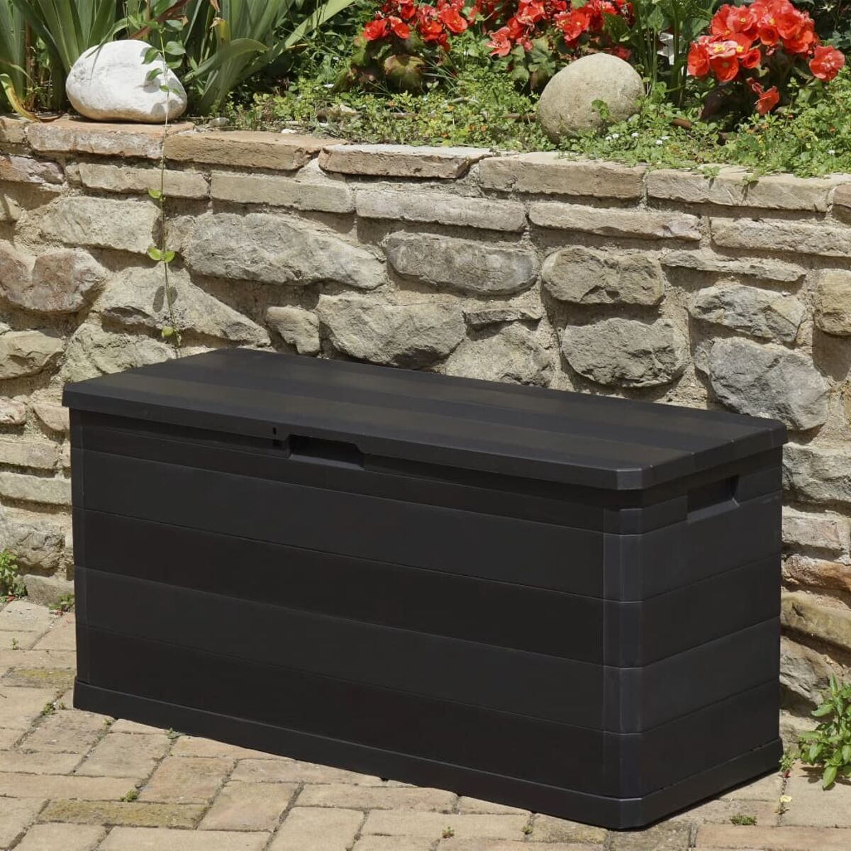 Eine schwarze Aufbewahrungsbox auf einer Backstein-Terrasse, mit Steinmauer und roten Blumen.