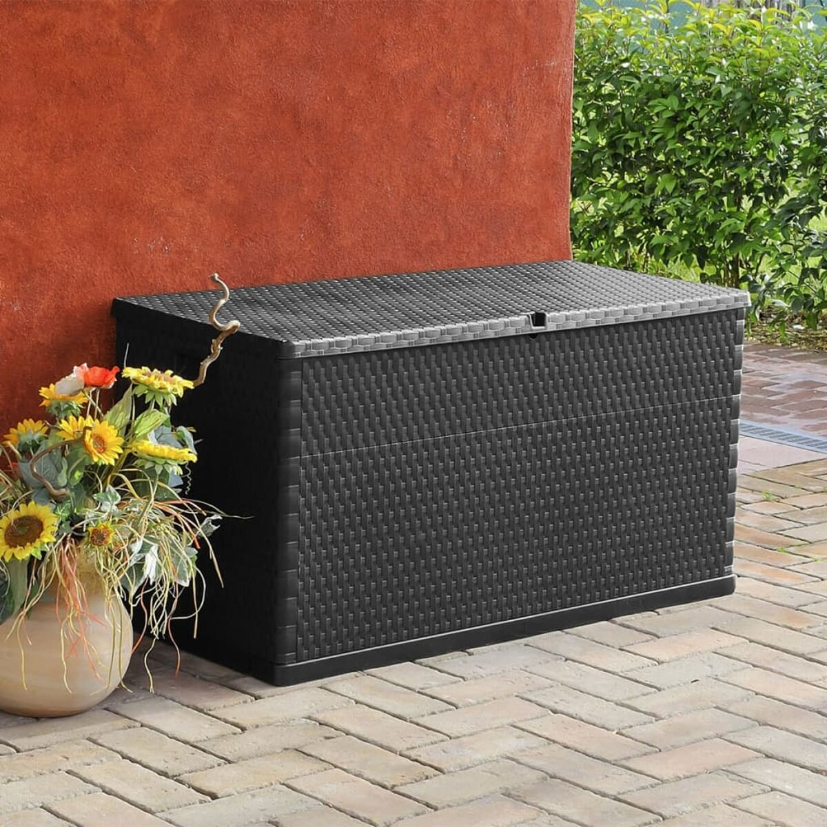 Schwarze Aufbewahrungsbox aus Rattan mit Deckel, neben Topfsonnenblumen auf Ziegelpflaster.