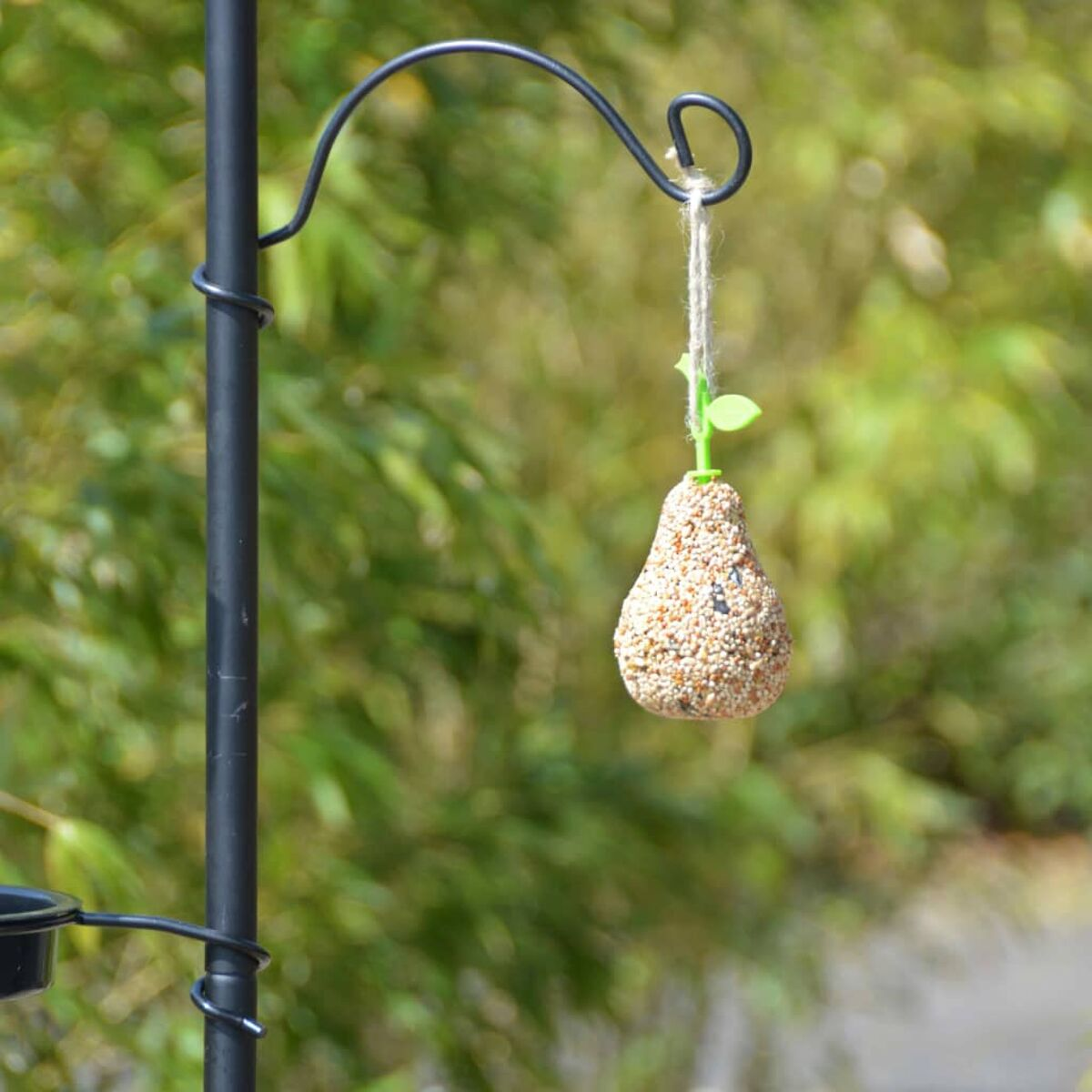 Vogelfutterspender in Birnenform, hängend an einem schwarzen Metallpfahl, verschwommener grüner Hintergrund.