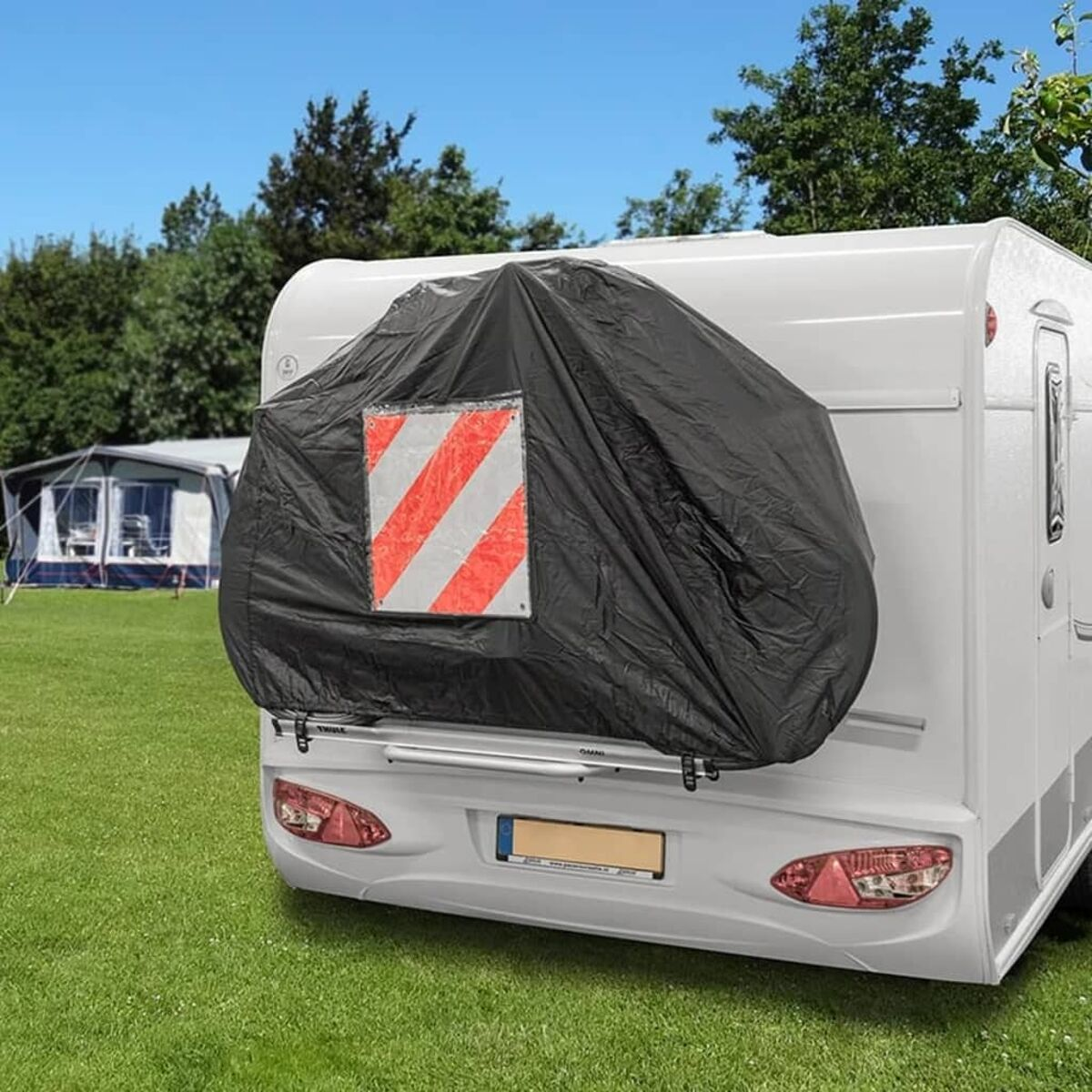 Weißer Wohnwagen mit schwarzer Abdeckung über einem Reifen, geparkt auf Gras. Bäume und Zelte im Hintergrund.