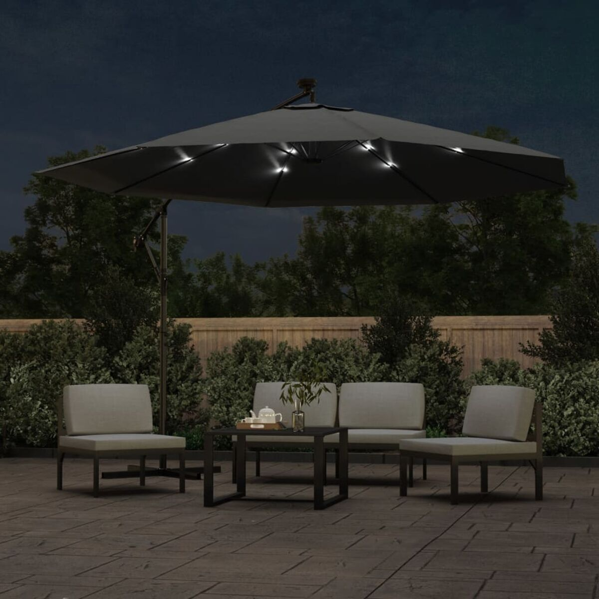 Außenszene auf der Terrasse bei Nacht. Grauer Schirm, Stühle und Tisch sind beleuchtet. Grüne Büsche.