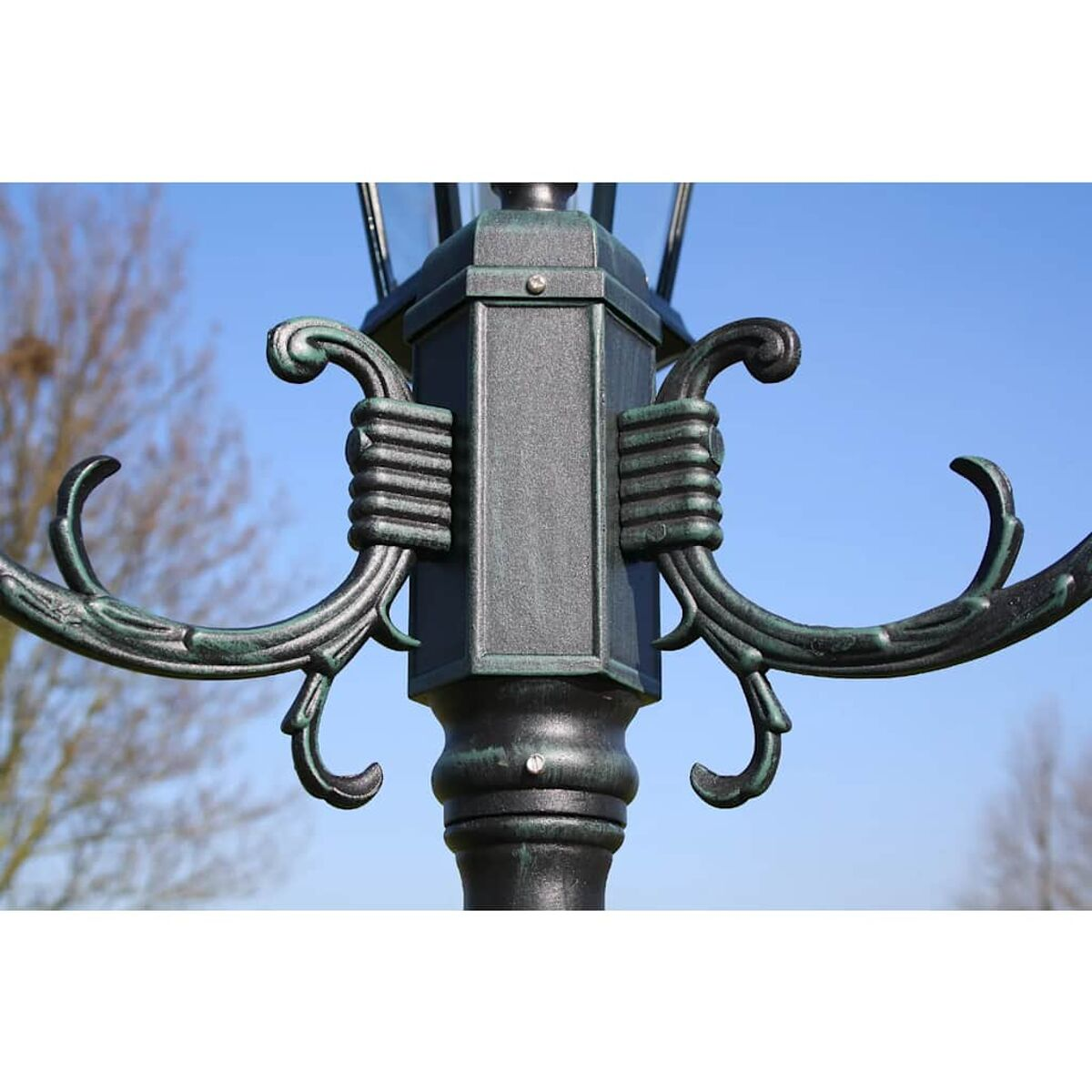 Nahaufnahme einer schwarz-grünen Straßenlaterne mit aufwendigen Details vor blauem Himmel und Bäumen.