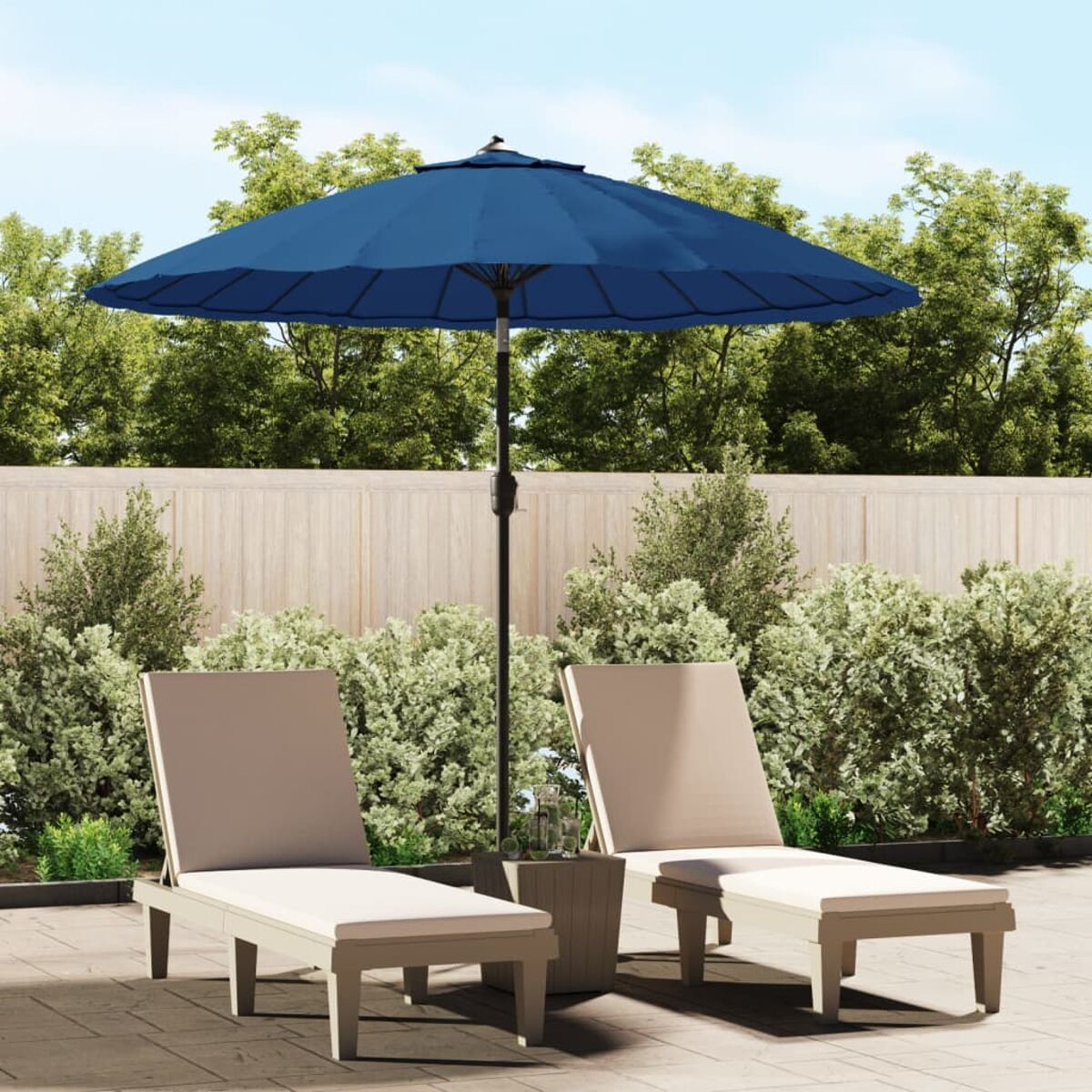 Zwei beige Liegestühle, ein blauer Schirm und ein kleiner Tisch auf der Terrasse. Grüne Sträucher und Bäume im Hintergrund.