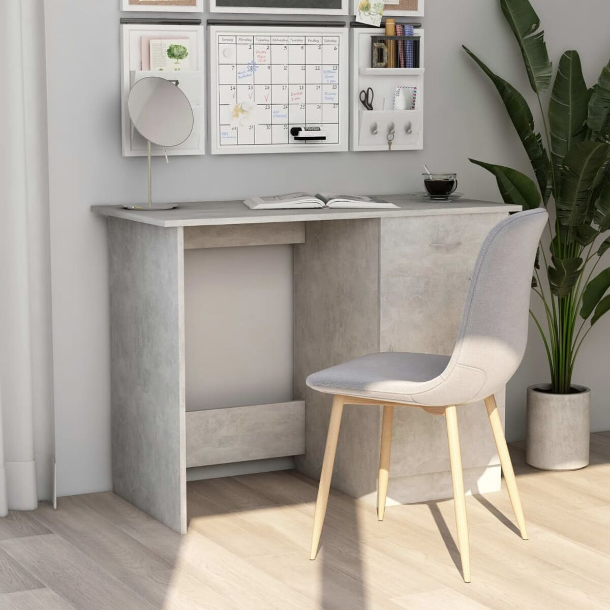 Ein grauer Schreibtisch mit Stuhl. An der Wand befinden sich ein Kalender, ein Spiegel und andere Gegenstände. Der Stuhl steht vor dem Schreibtisch.