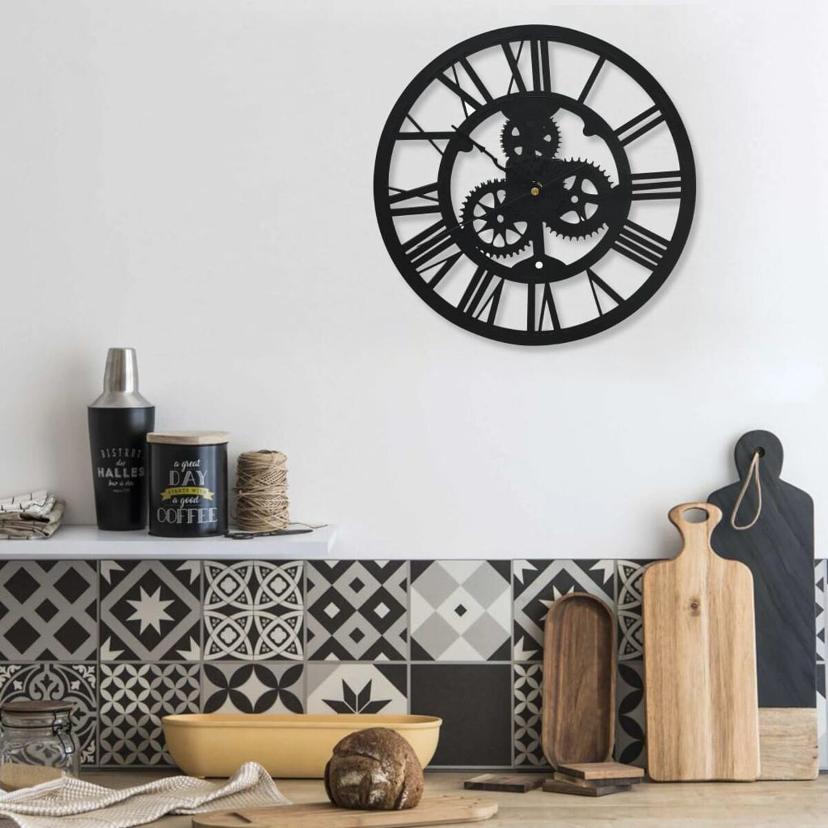 Orologio nero a ingranaggi con numeri romani su una parete bianca sopra un bancone della cucina con decorazioni.