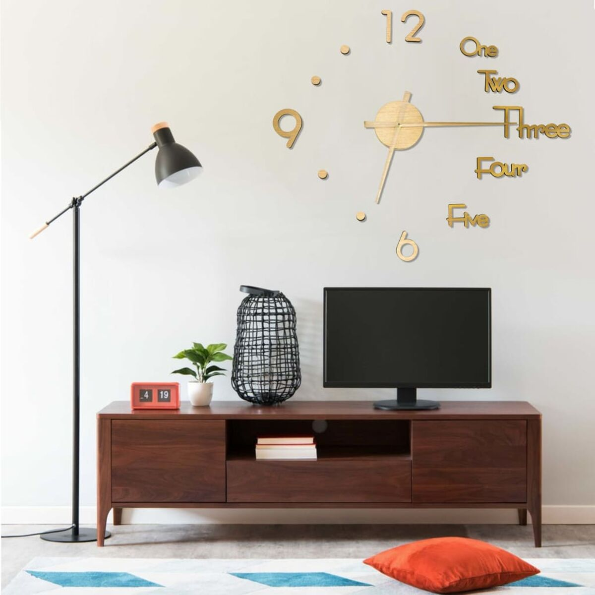 Wohnzimmer mit Wanduhr, Fernseher und Möbeln. Eine Lampe und ein rotes Kissen sind sichtbar.