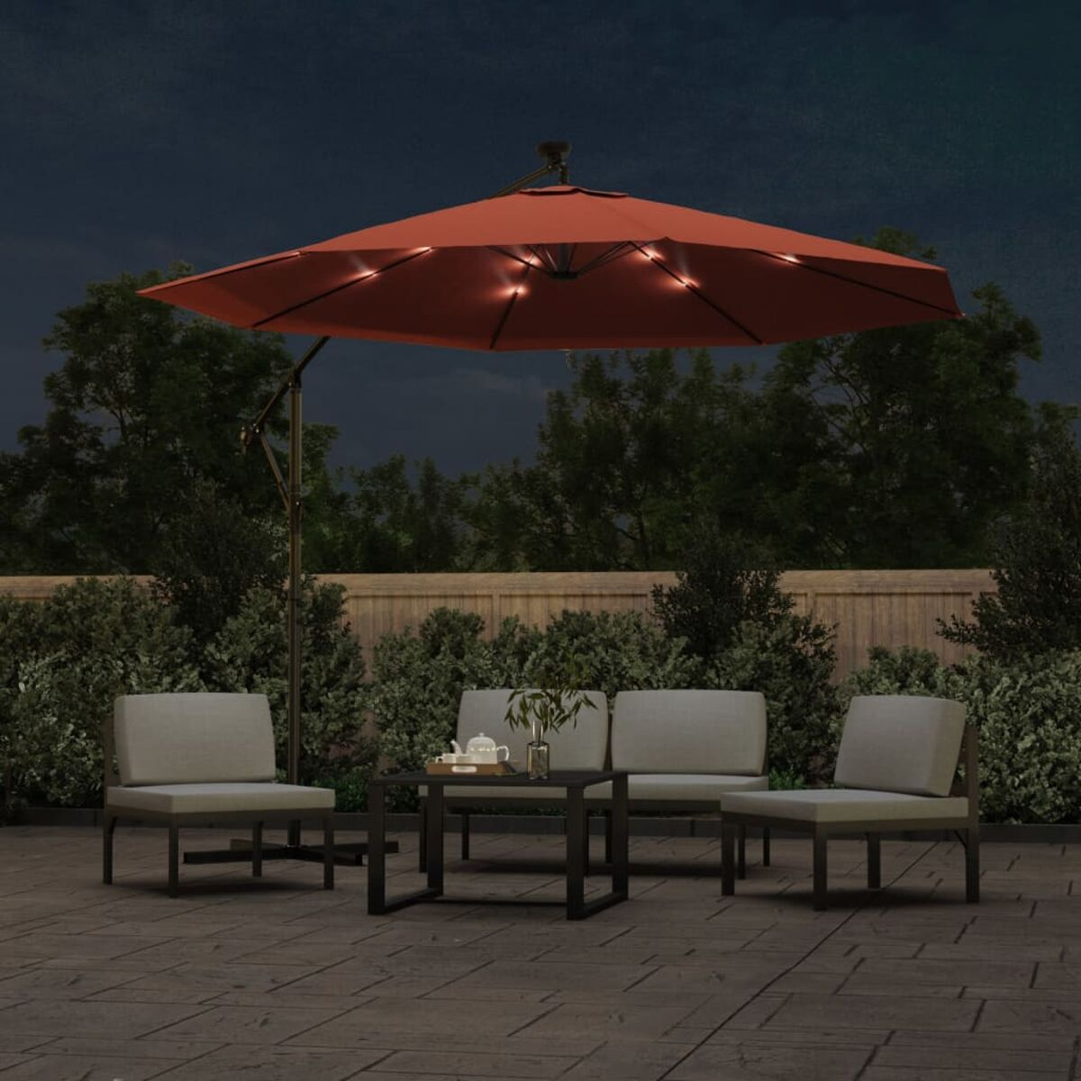 Terrassenszene bei Nacht: roter Schirm, vier Stühle und ein Tisch. Lichter sind an. Büsche im Hintergrund.