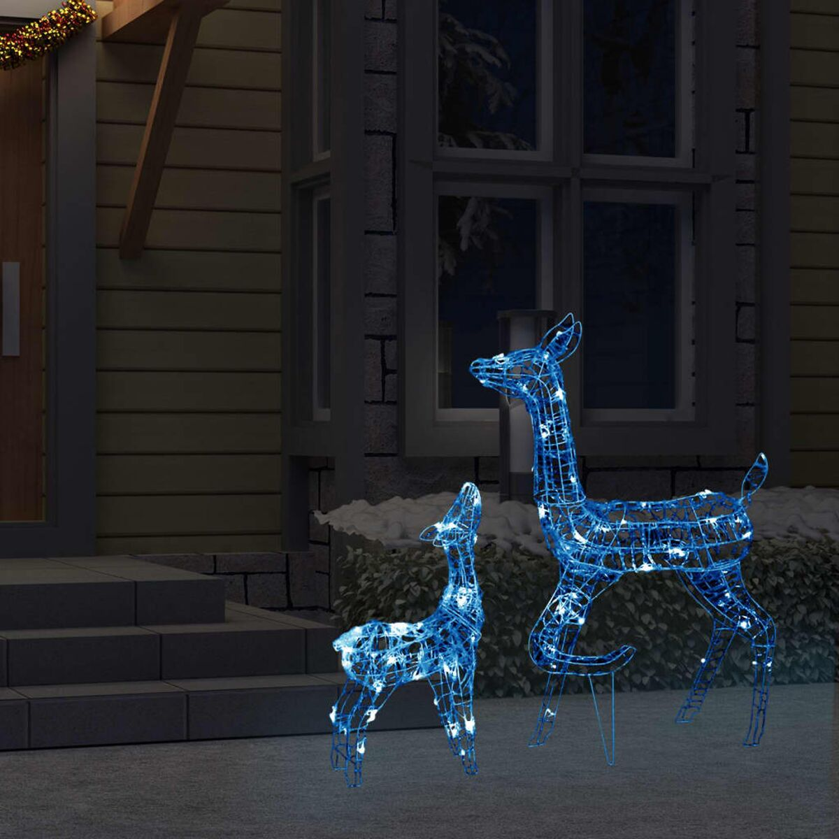 Zwei blau leuchtende Hirschdekorationen, eine große und eine kleine, vor einem Haus bei Nacht.