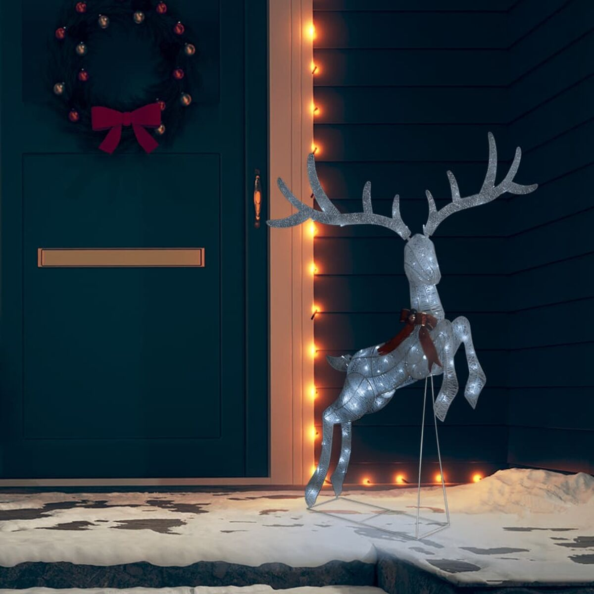 Silberne Rentier-Weihnachtsdekoration mit Lichtern, auf Schnee vor einer Tür.