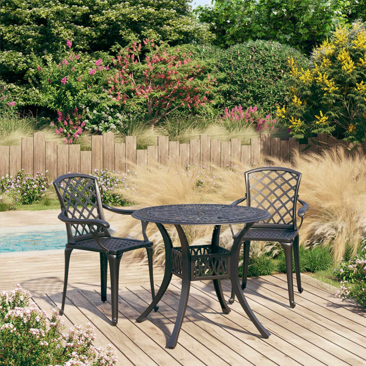Ein dunkler Metalltisch und zwei Stühle auf einer Holzterrasse. Grüne Büsche und Blumen im Hintergrund.
