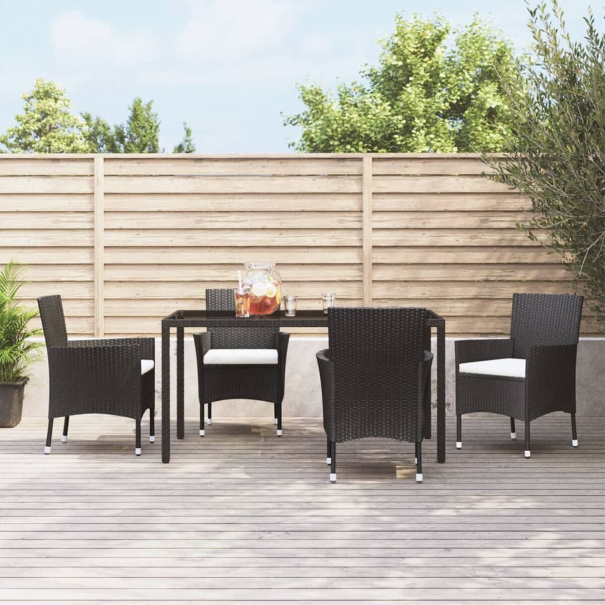 Essgruppe für draußen. Enthält einen Tisch, vier Stühle und eine Glaskanne. Holzterrasse und Zaun.