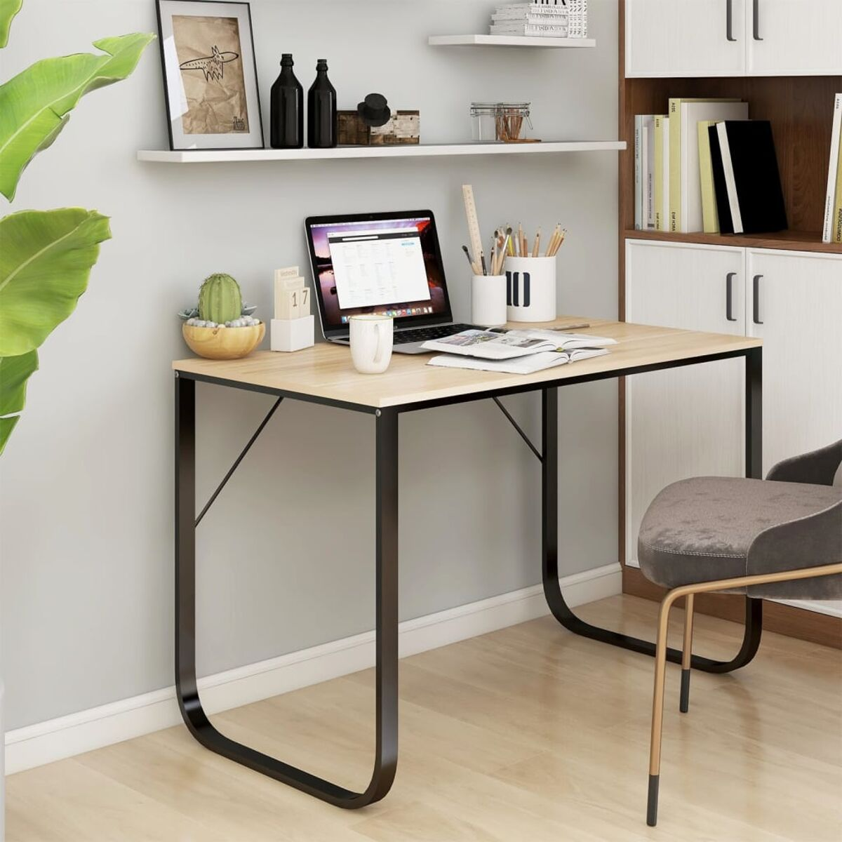Kleiner Schreibtisch mit Laptop, Büchern, Bleistiften und einem Kaktus. Weiße Wand und Regale im Hintergrund.