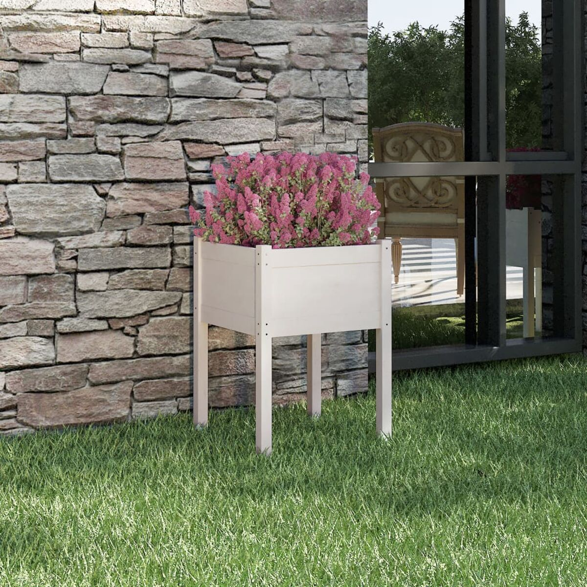 Weiße Pflanzkasten mit rosa Blumen, auf grünem Gras neben einer Steinmauer.