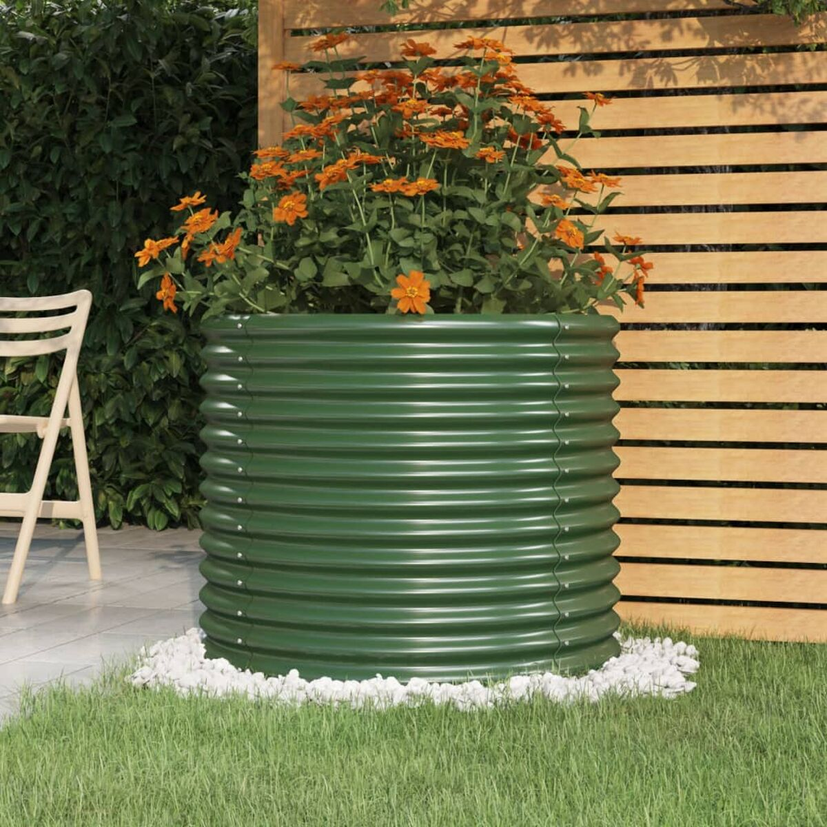 Grüner Metallpflanzer mit orangefarbenen Blumen. Weiße Steine umgeben die Basis, mit Gras und Zaun.