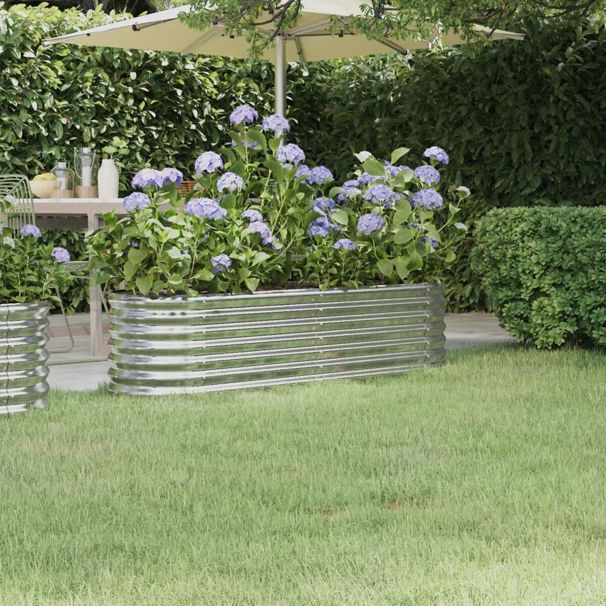 Metall-Gartenpflanzer mit blauen Blumen und grünem Gras. Hintergrund: Schirm und Sträucher.