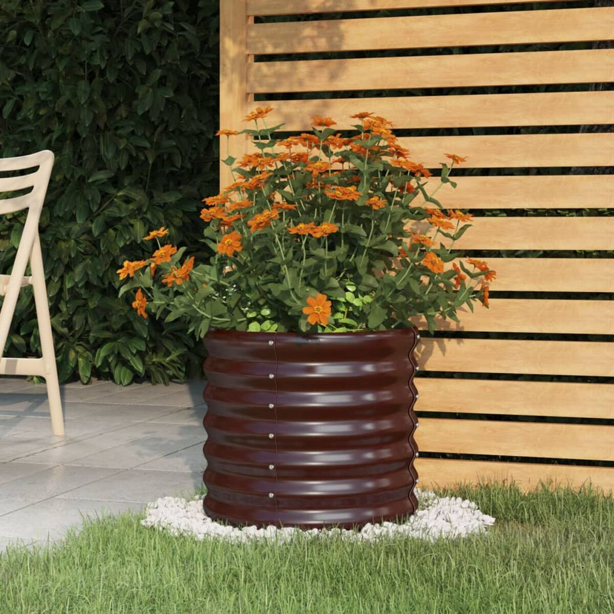 Dunkelbrauner, gerippter Metallblumentopf mit orangen Blumen. Weiße Kiesel umgeben ihn. Holzzaun.