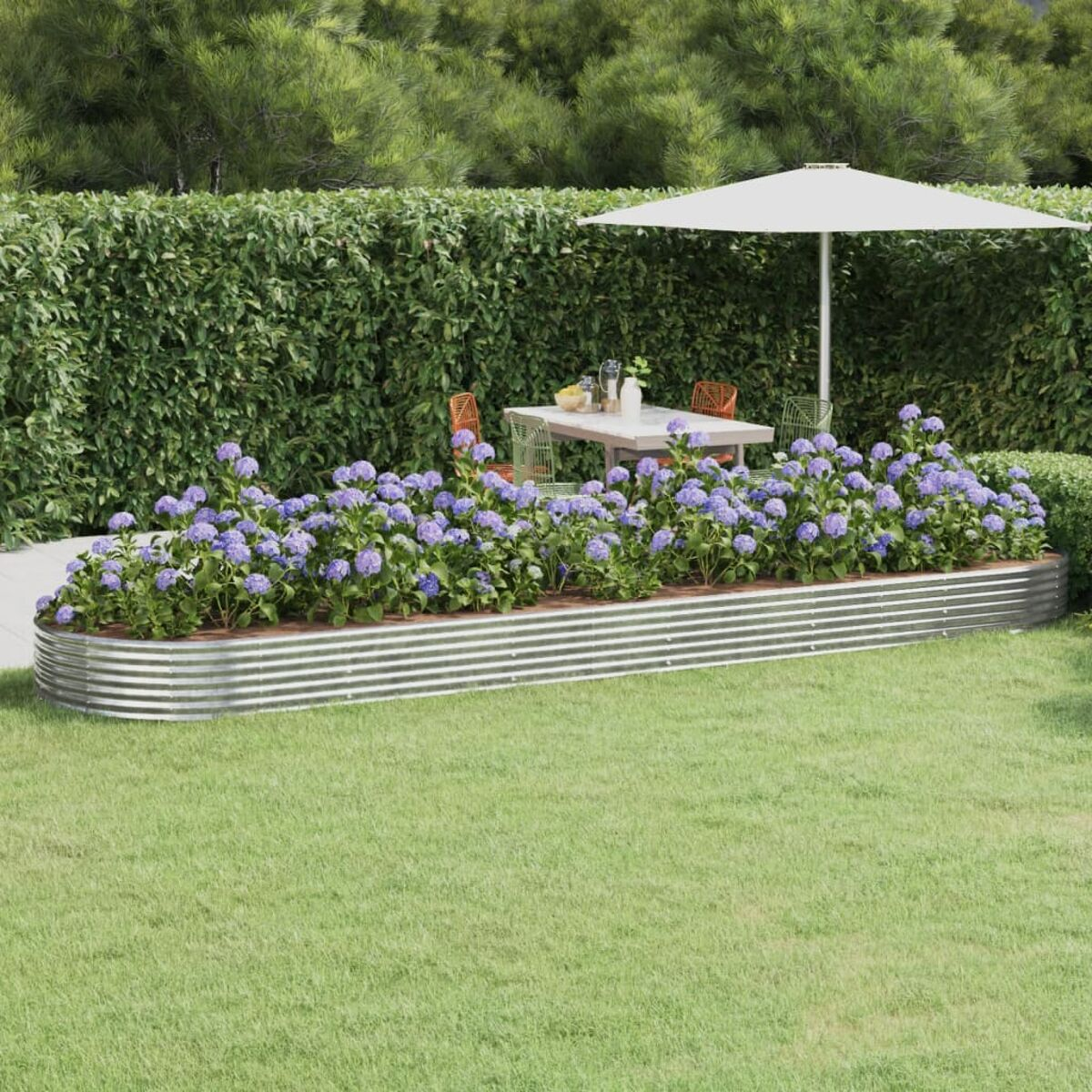Ein erhöhtes Gartenbeet aus Metall mit blauen Blumen, ein weißer Sonnenschirm, ein Tisch und eine grüne Hecke im Hintergrund.