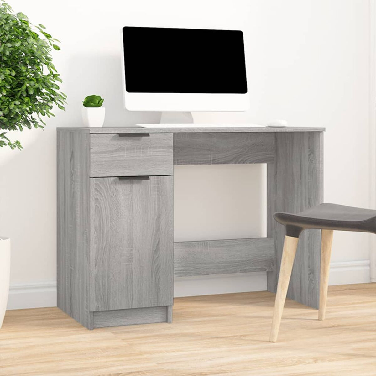 Ein grauer Schreibtisch mit Computer, Pflanze und Stuhl vor weißer Wand.