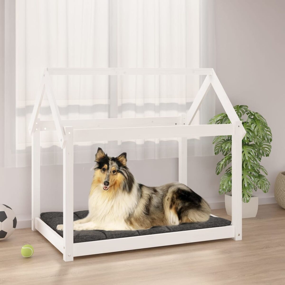 Ein Hund liegt in einem weißen Hundebett aus Holz. Das Bett ist hausförmig. Ein Ball liegt auf dem Boden.