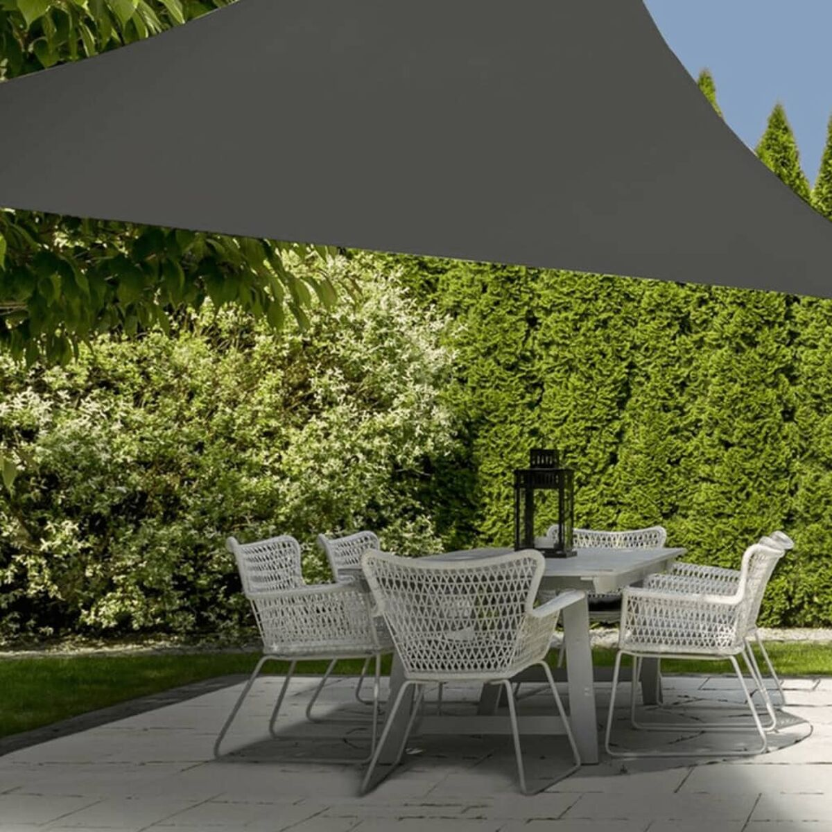 Eine Hinterhofszene mit einer weißen Terrasse, weißen Stühlen um einen Tisch und einem großen, dunkelgrauen Sonnensegel darüber.