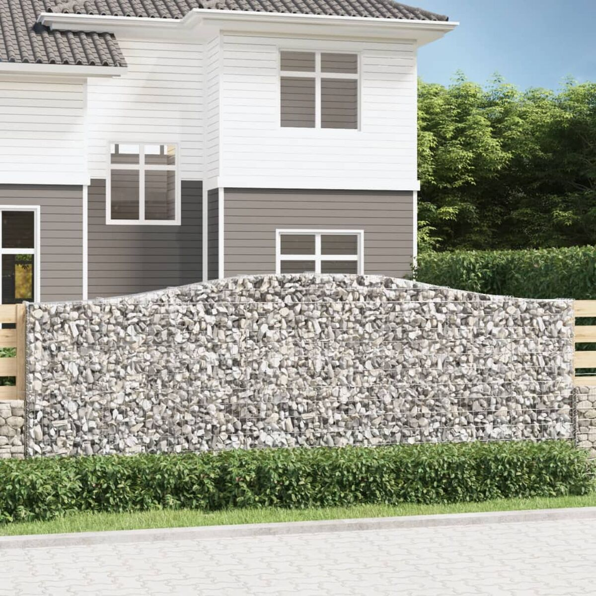 Ein Haus mit einer Steingabionenmauer und einem Holzzaun, mit einer grünen Hecke und blauem Himmel.